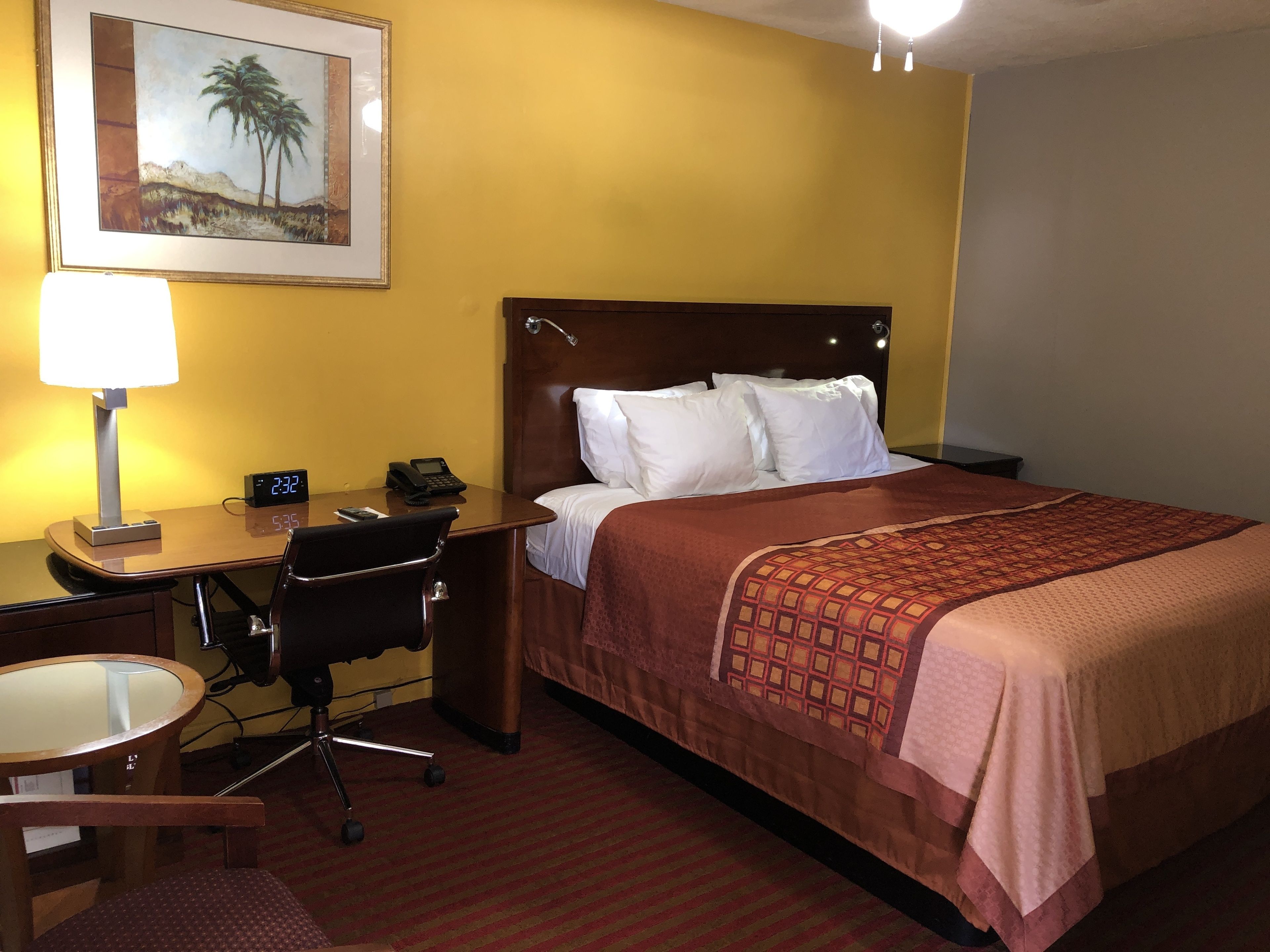 A hotel room with a bed, a desk with a lamp, a chair, a clock, and a framed picture on the wall.
