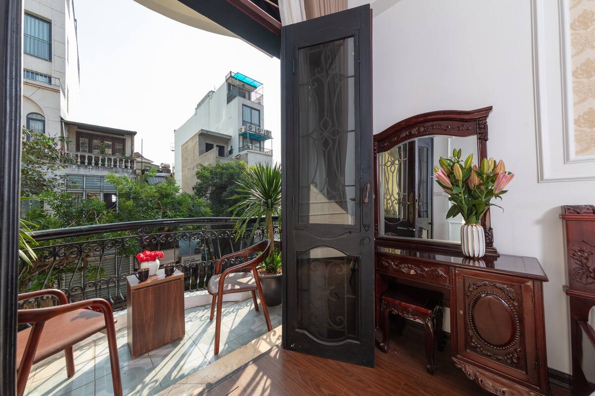A room with a balcony, a wooden desk, a chair, a mirror, and a vase with flowers.