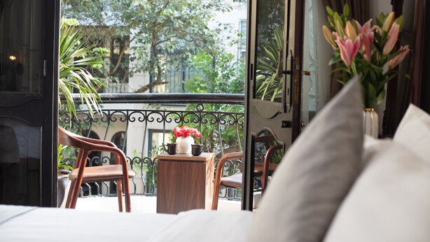 Une chambre d’hôtel avec un balcon, une chaise en bois, une petite table et un vase rempli de fleurs.