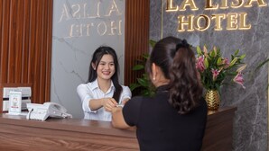 Un comptoir d’accueil d’hôtel avec un réceptionniste et un client. Le nom de l’hôtel, « LA LISA HOTEL », est visible sur le mur.