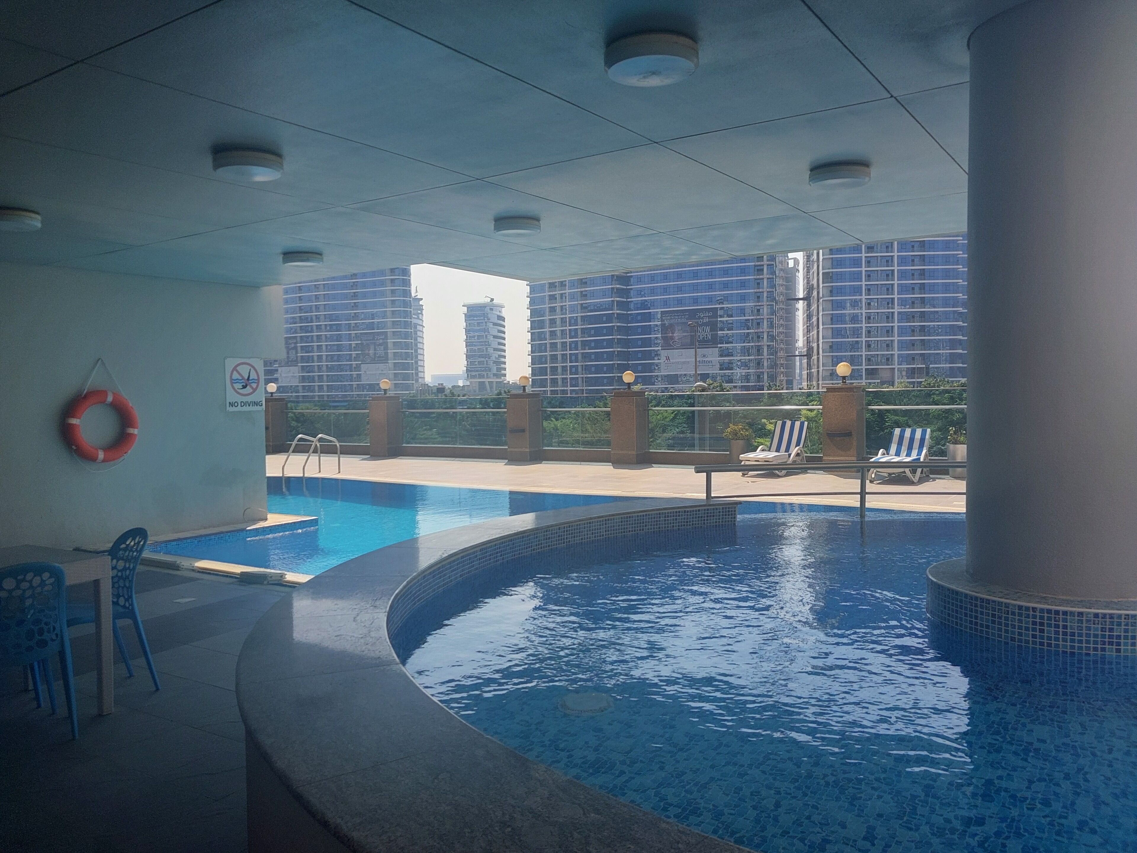 Une piscine intérieure avec un panneau indiquant qu’il est interdit de plonger, des chaises longues et une vue sur les immeubles de grande hauteur.