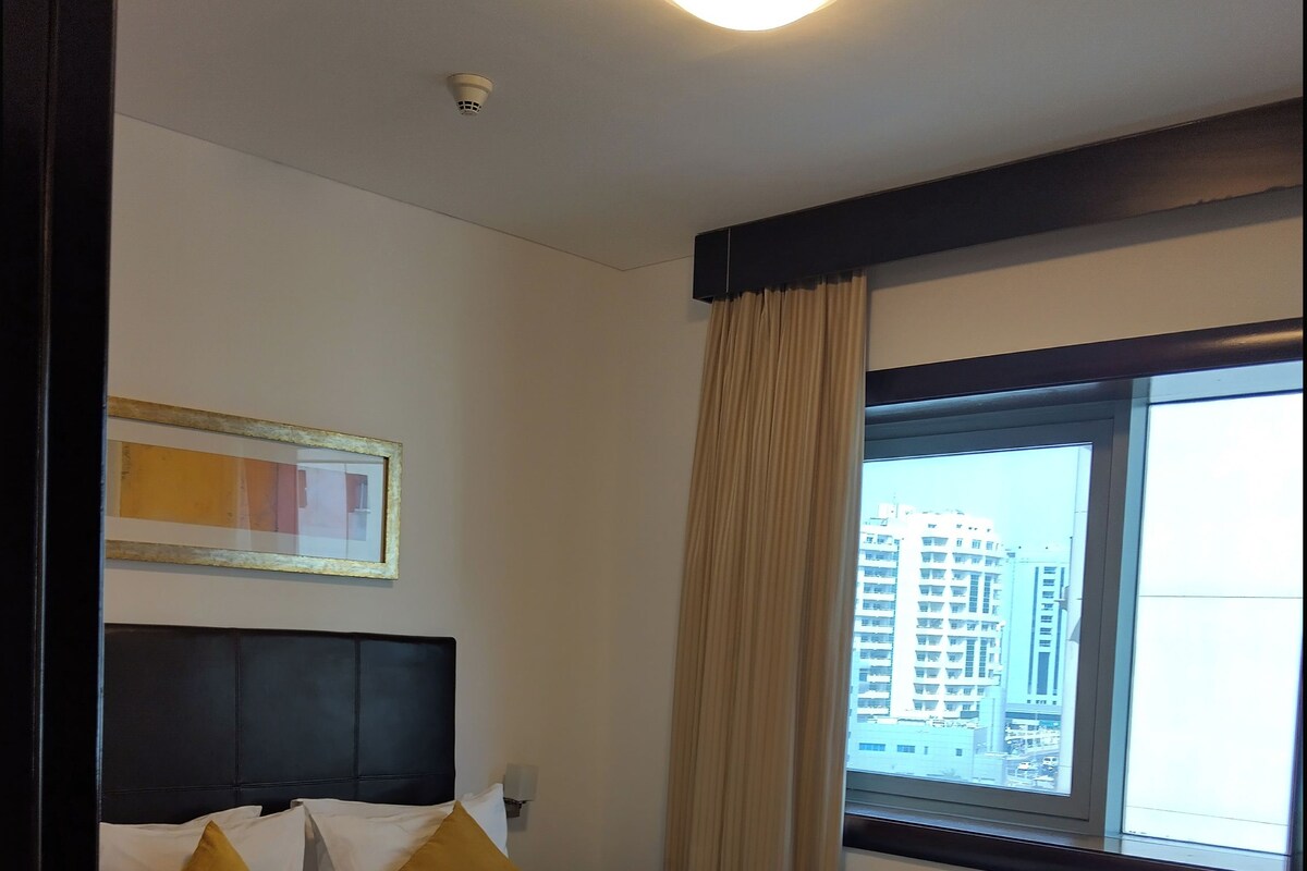 Une chambre d’hôtel avec un lit, une tête de lit, une table de chevet, une fenêtre donnant sur des immeubles et un luminaire au plafond.