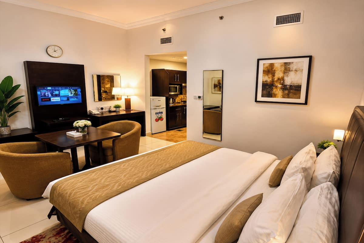 A hotel room with a bed, a desk with a chair, a television, a kitchen area, and a dining table.