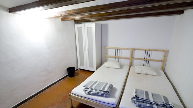 Alojamiento pequeño de una sola habitación con dos camas individuales, cabecero de madera, armario blanco y suelo de baldosas.