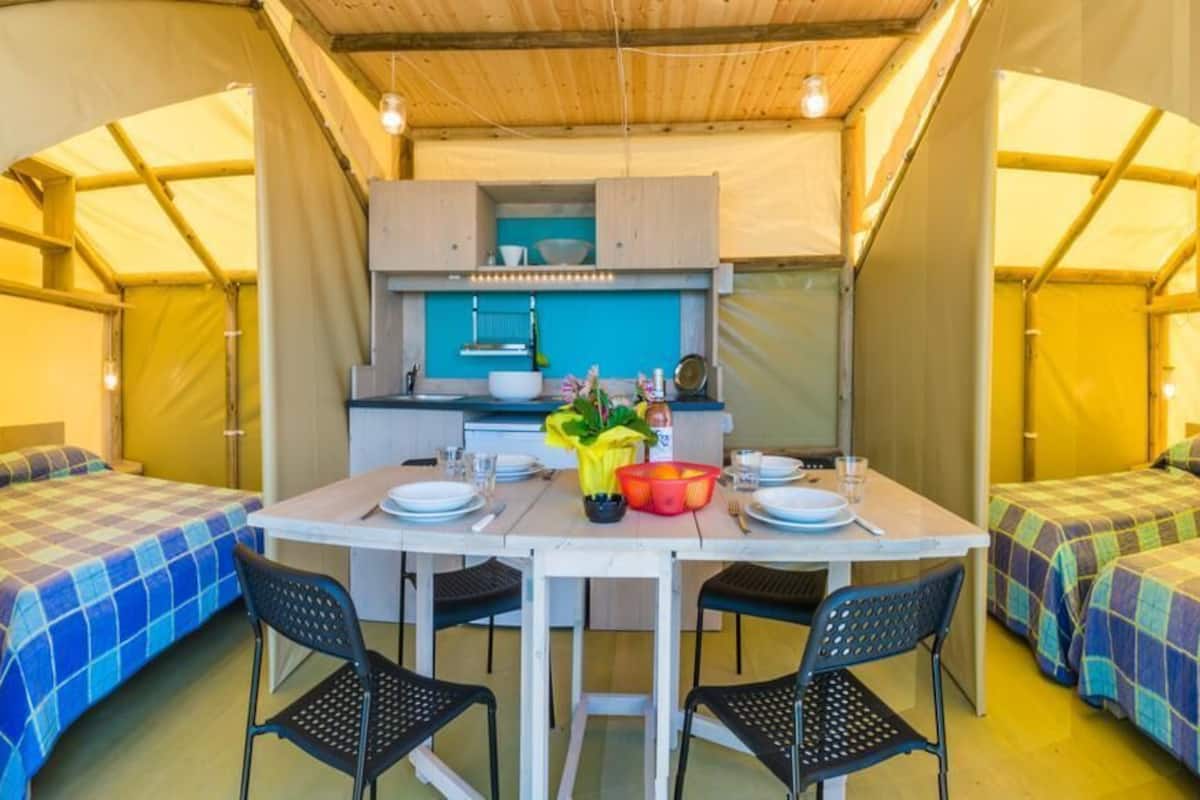 A tented accommodation with a central dining table, four black chairs, and two single beds with checkered bedding.