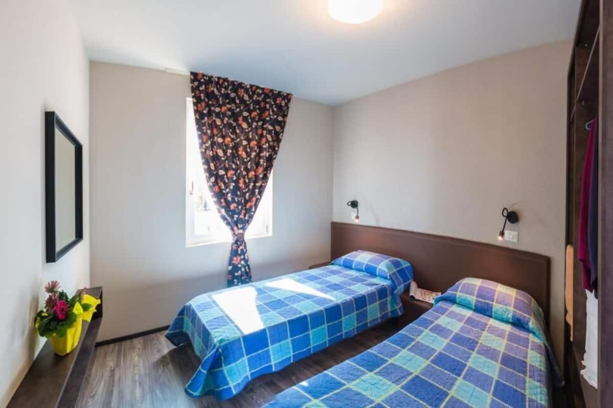 A small, clean room with two beds, a window with a floral curtain, a bedside table with a lamp, and a framed picture on the wall.