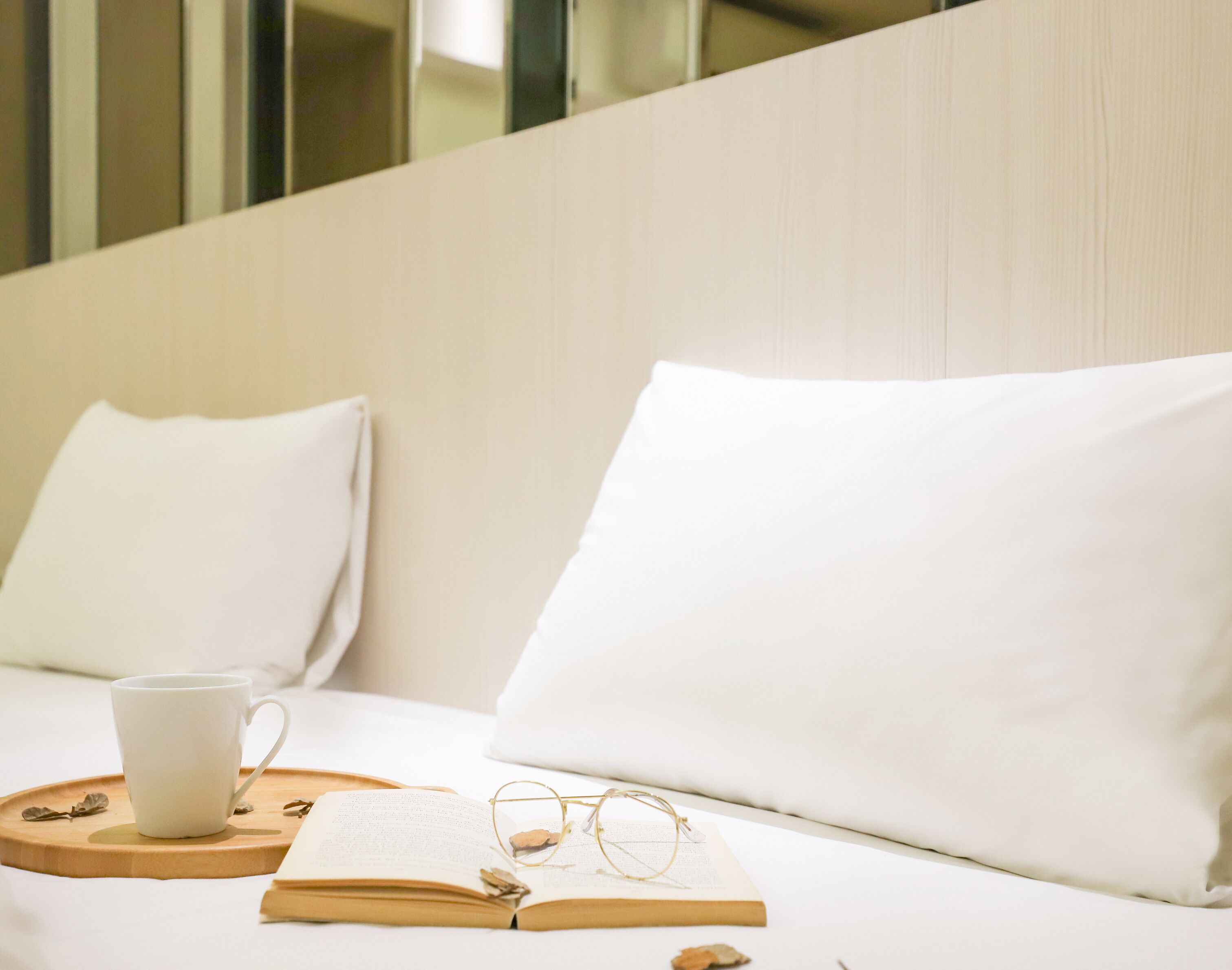 A neatly made bed with white linens, a white cup, a pair of glasses, and an open book on a wooden tray.