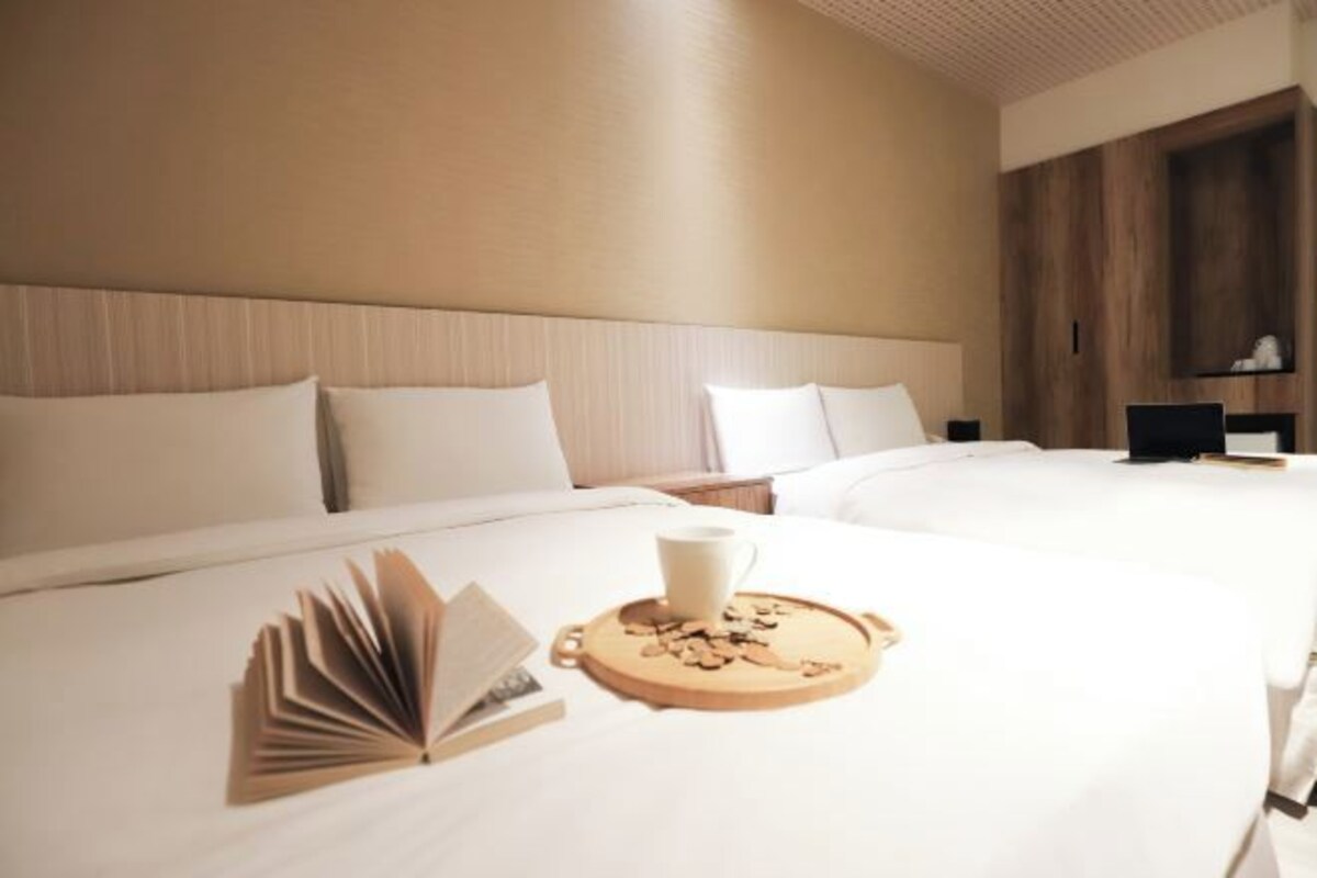 Ein Hotelzimmer mit zwei Betten, einem hölzernen Kopfteil, einem Nachttisch mit Lampe und einem Tablett mit Buch und Tasse.