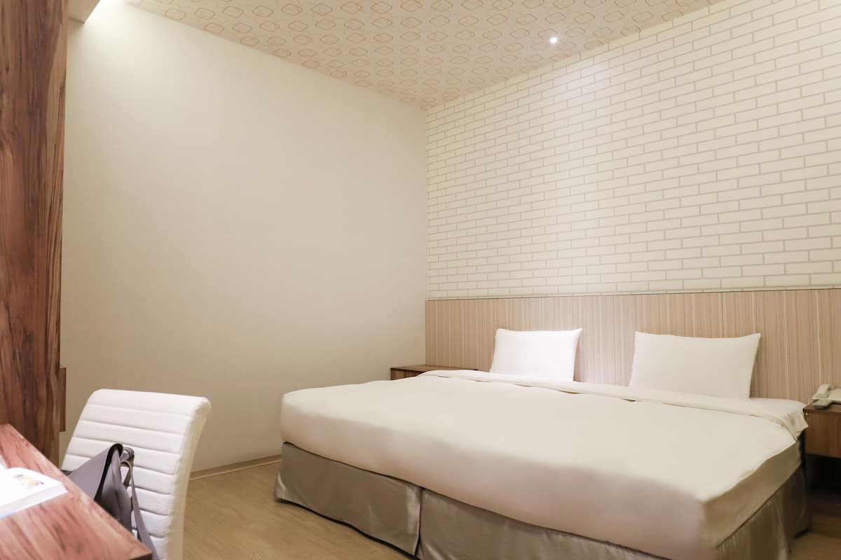 A modern bedroom with a wooden headboard, a large bed with white bedding, a desk with a chair, and a patterned ceiling.