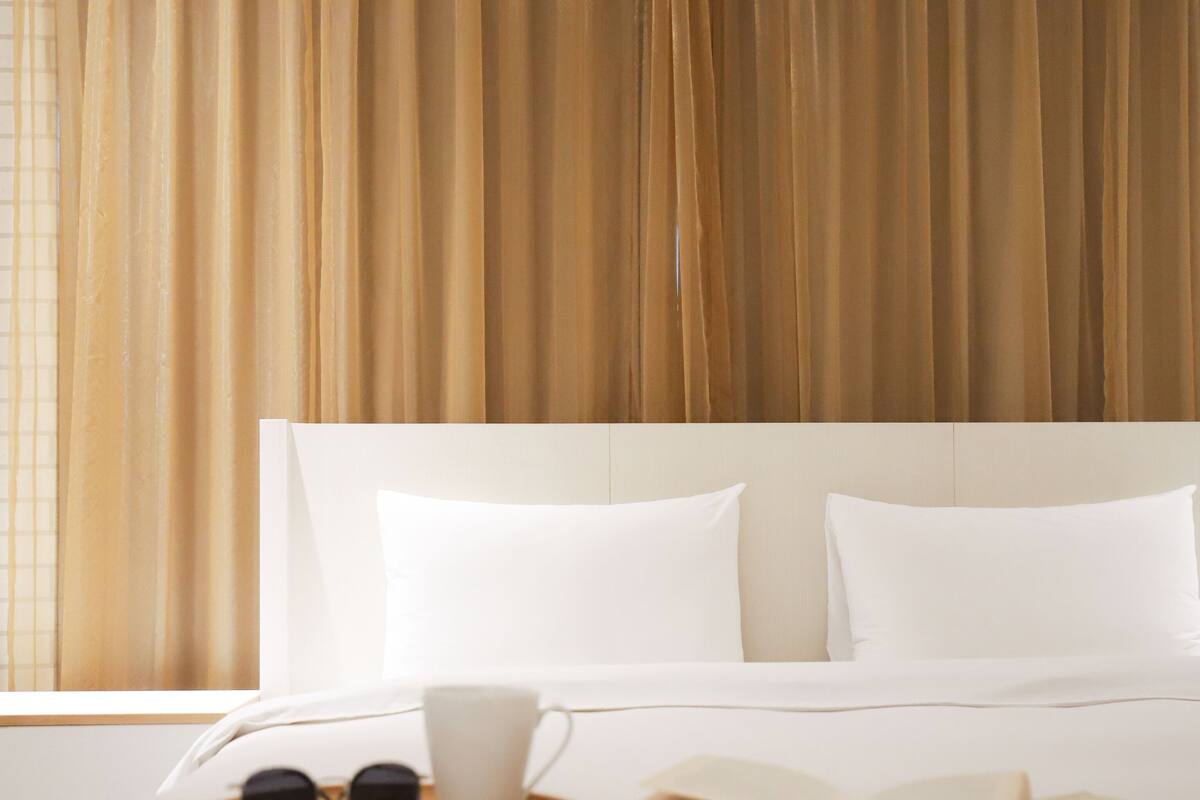 A neatly made bed with white linens, a pair of sunglasses, a white cup, and an open book on a wooden tray.