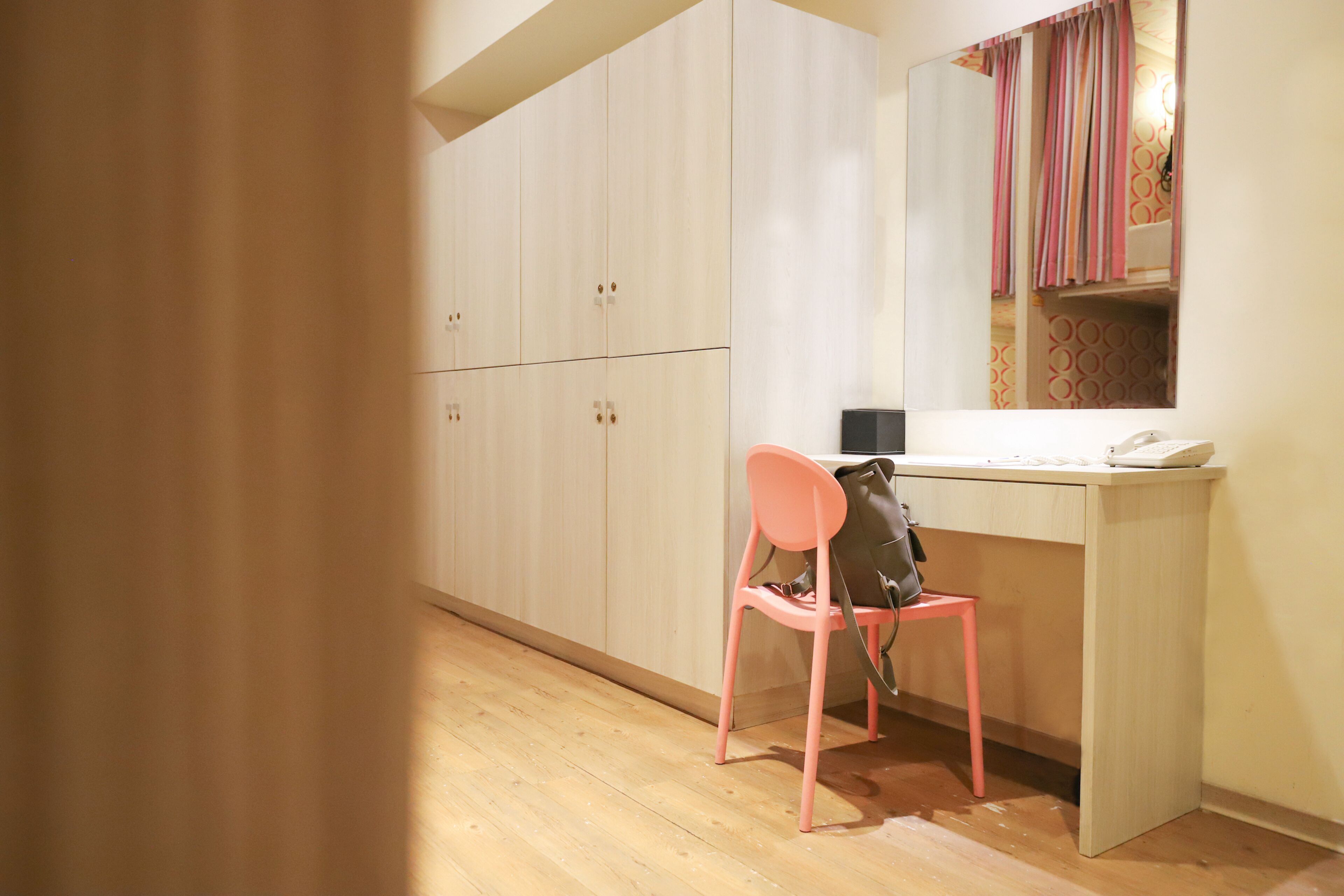 A room with a wooden floor, a pink chair, a desk with a computer, and a mirror reflecting a patterned curtain.