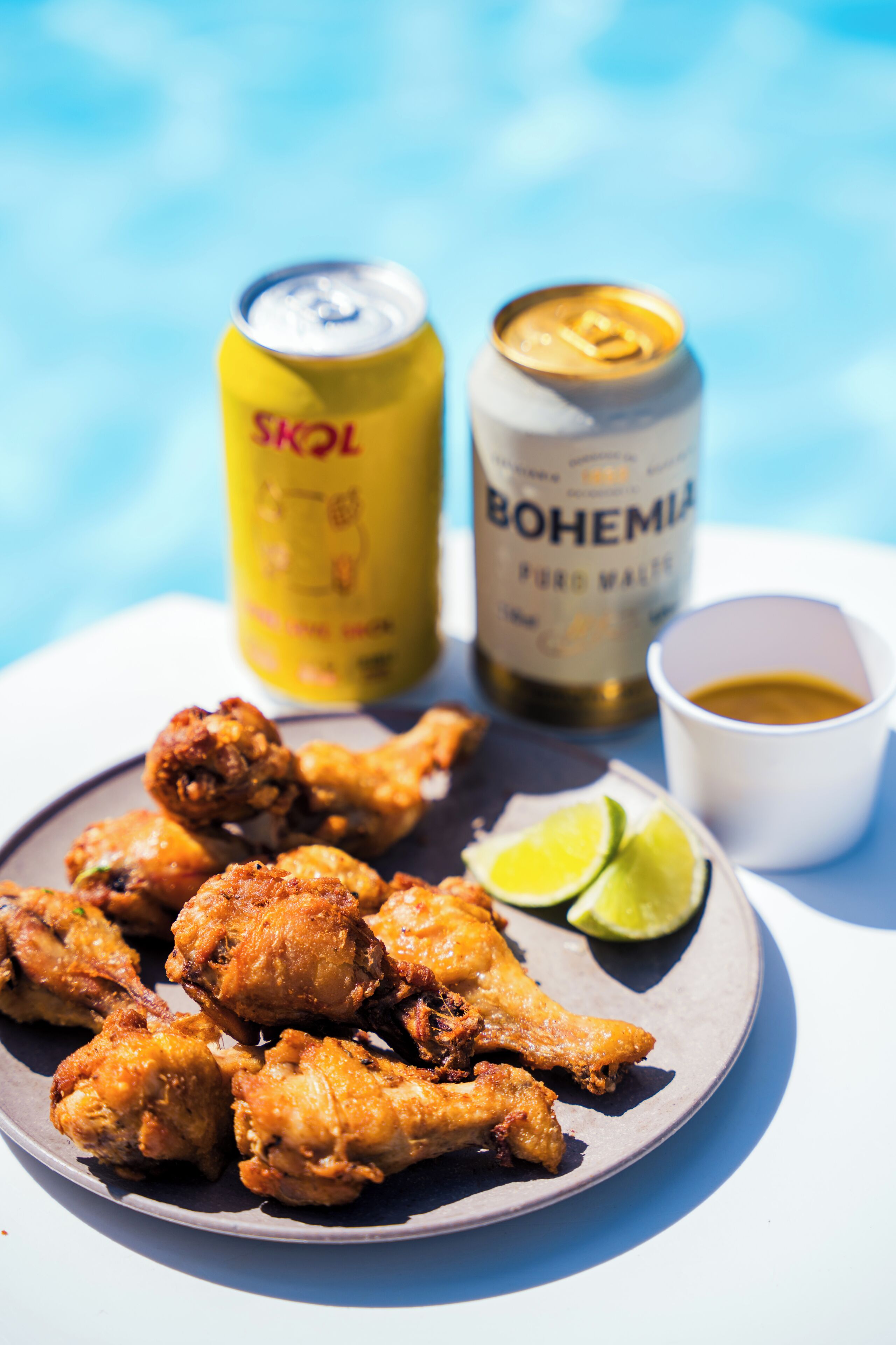 A plate of fried chicken wings with lime wedges and a small cup of sauce, a can of beer, and a bottle of beer.