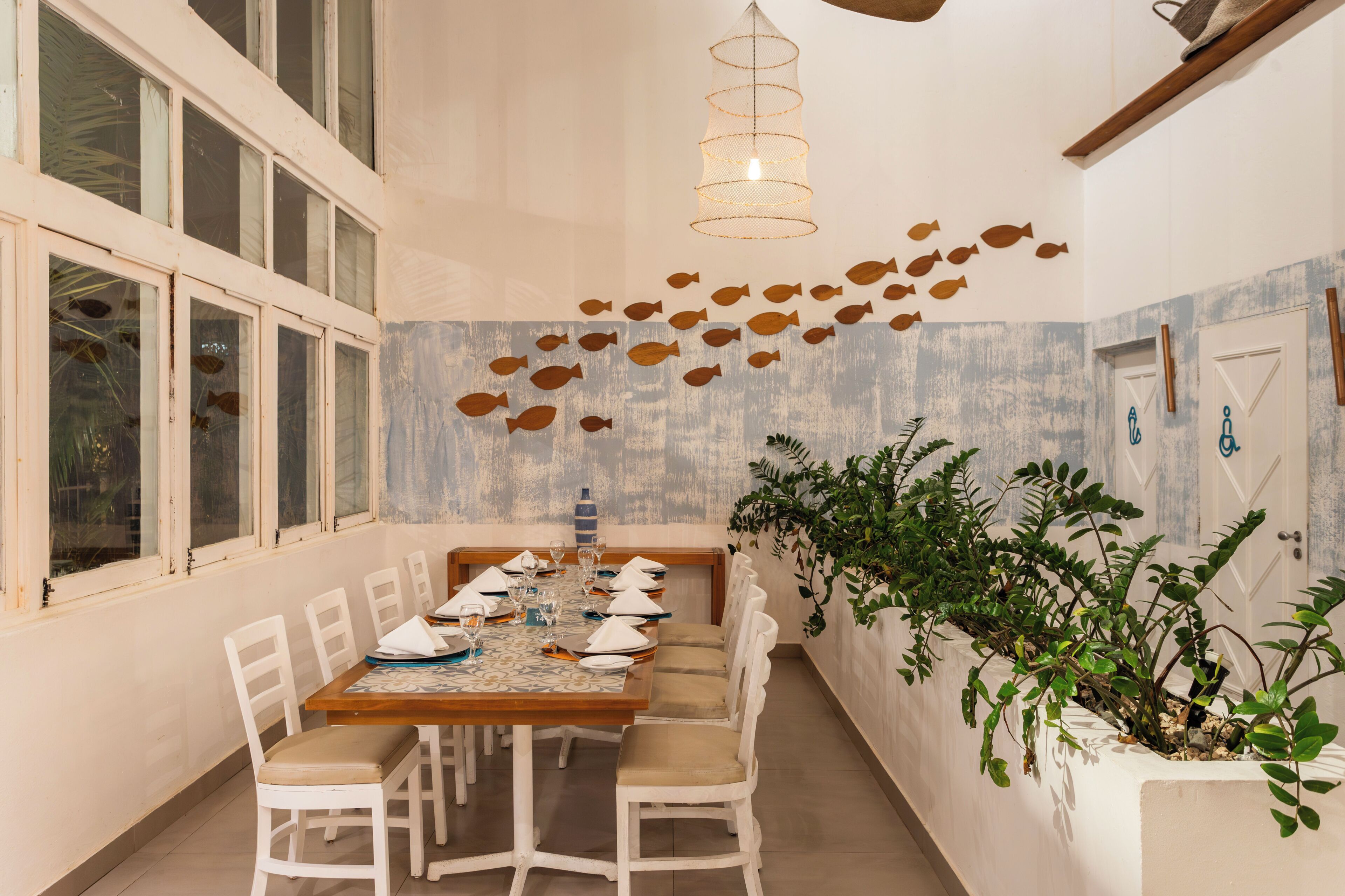 Un coin repas avec une table en bois dressée pour six personnes, des chaises blanches, un luminaire suspendu et un mur décoré de motifs de poissons.