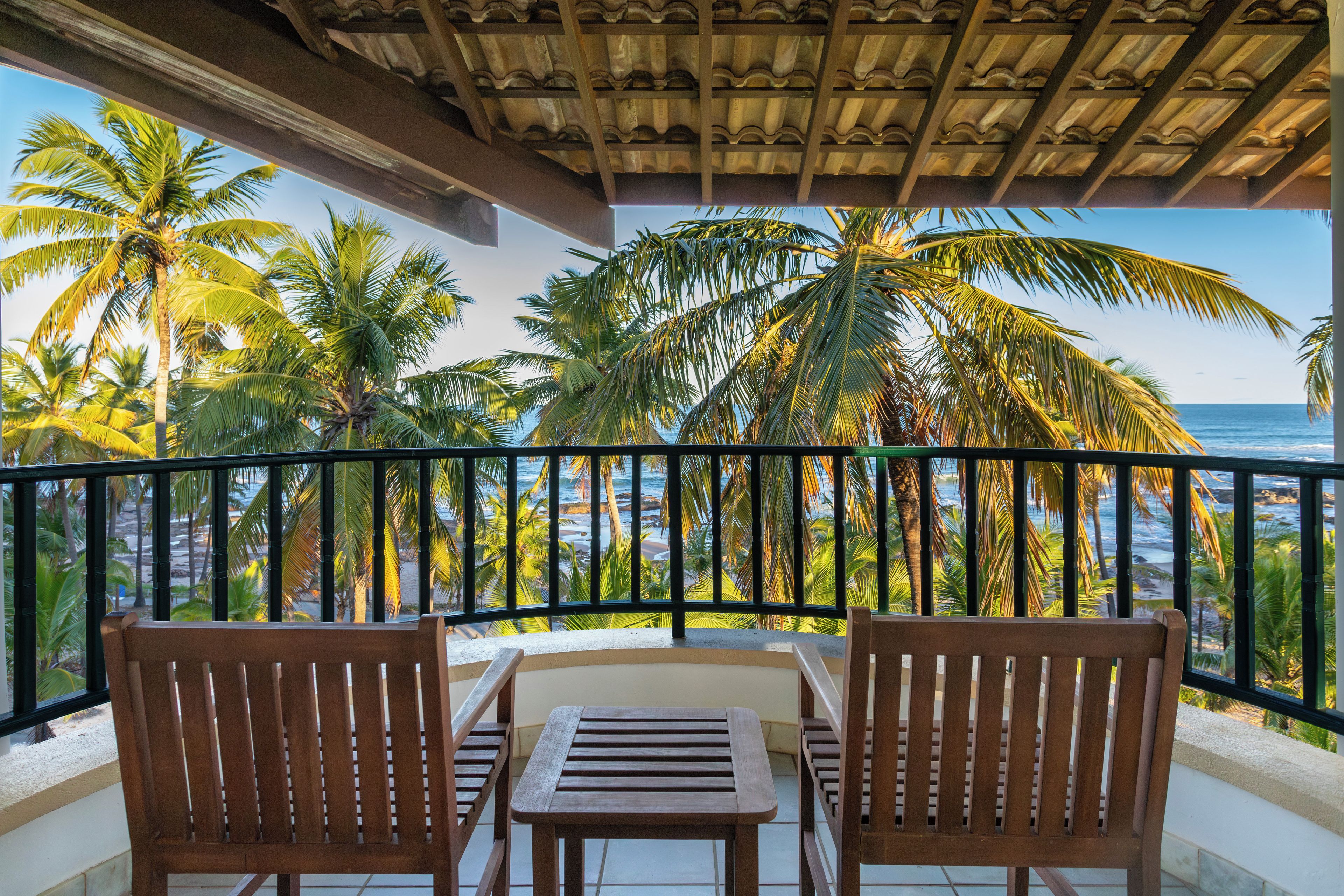 A balcony with two wooden chairs and a table, overlooking a beach with palm trees and the ocean.