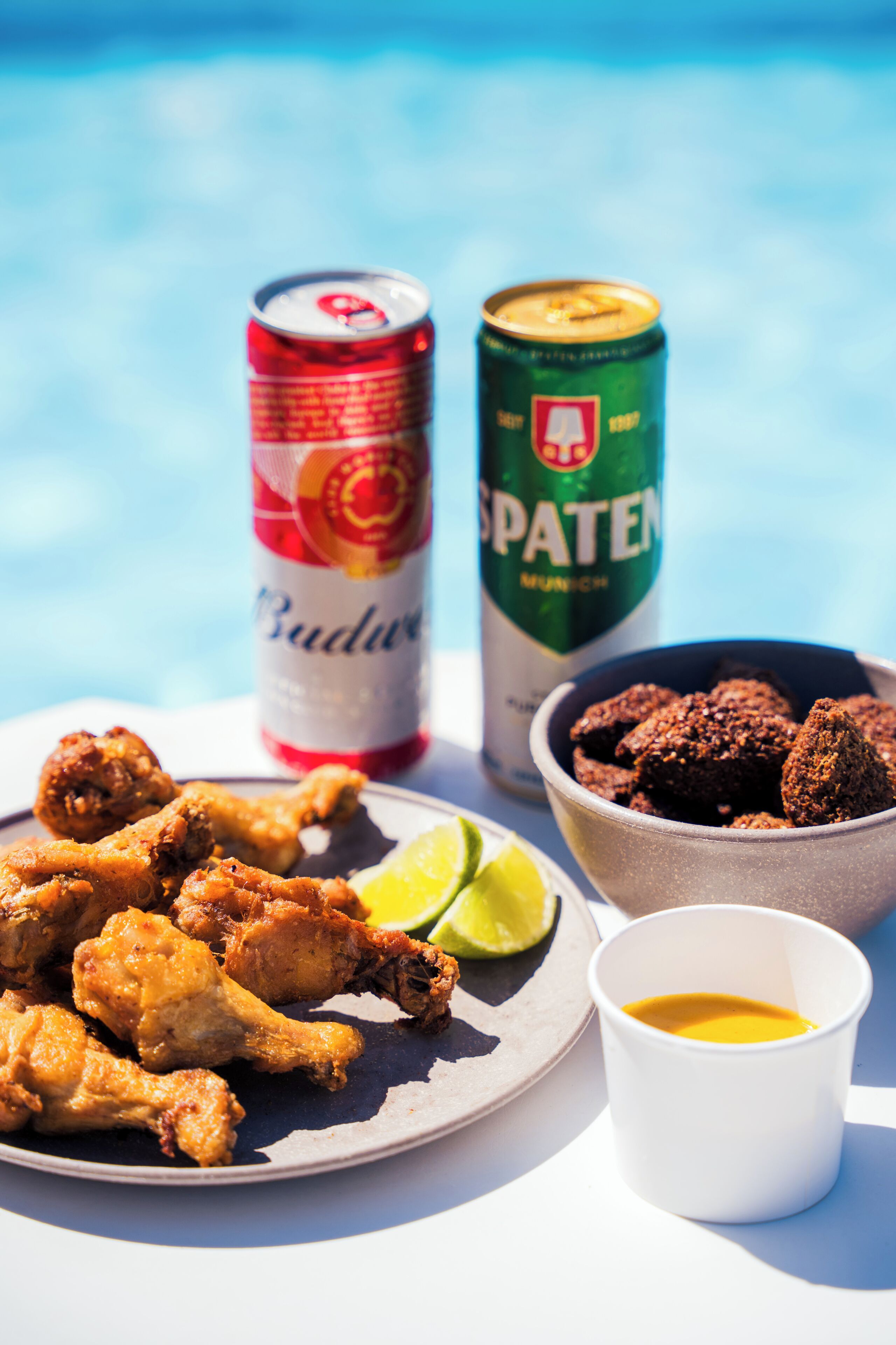 Een bord gefrituurd kippenvlees, een blikje Budweiser, een blikje Spaten, een schaaltje chocoladesnacks, een partje limoen en een klein bekertje saus.