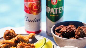 A plate of fried chicken, a can of Budweiser, a can of Spaten, a bowl of chocolate-covered snacks, a lime wedge, and a small cup of sauce.