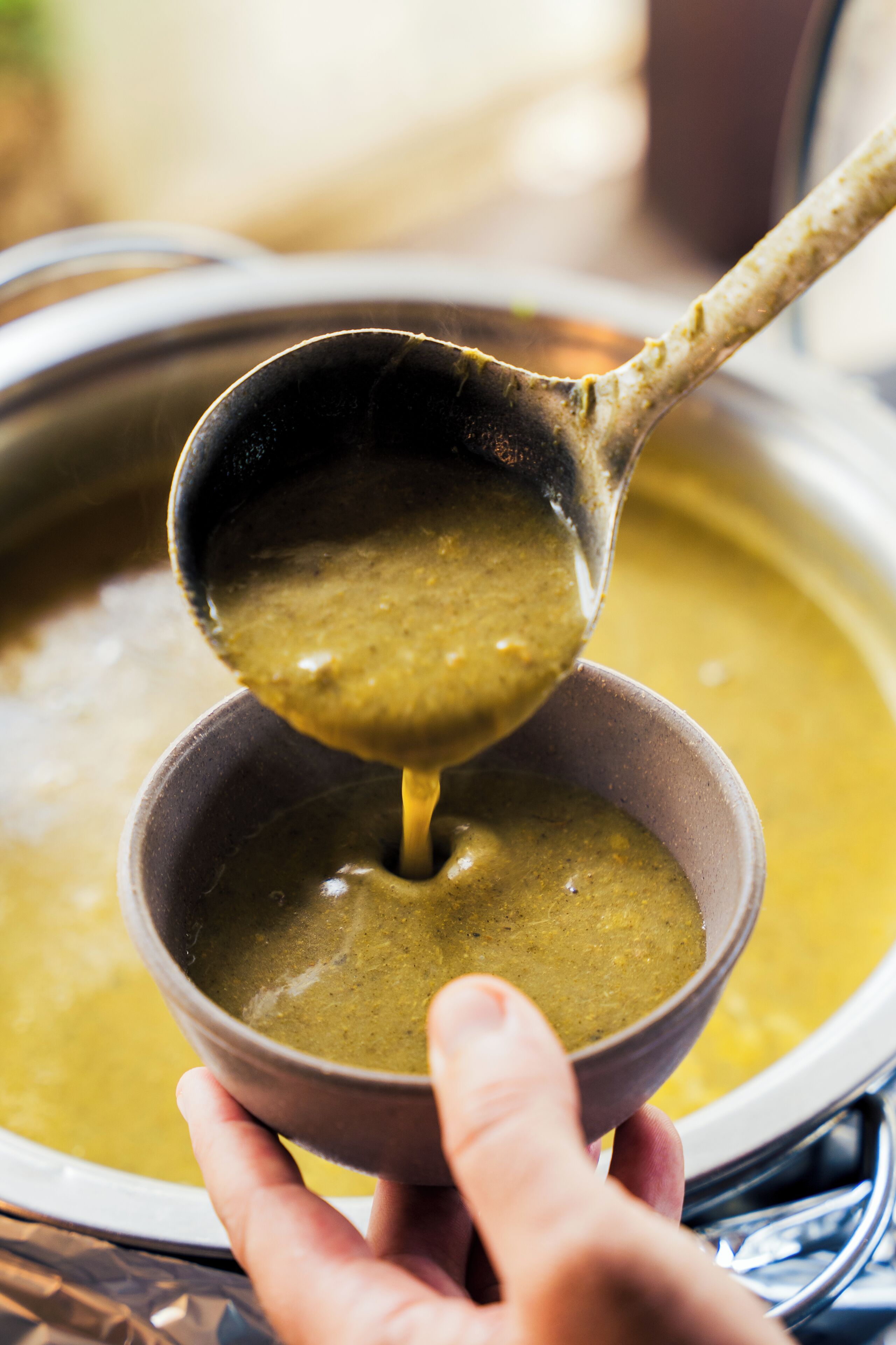 Une main tenant un bol dans lequel une louche verse une soupe épaisse et verdâtre, provenant d’une plus grande marmite.