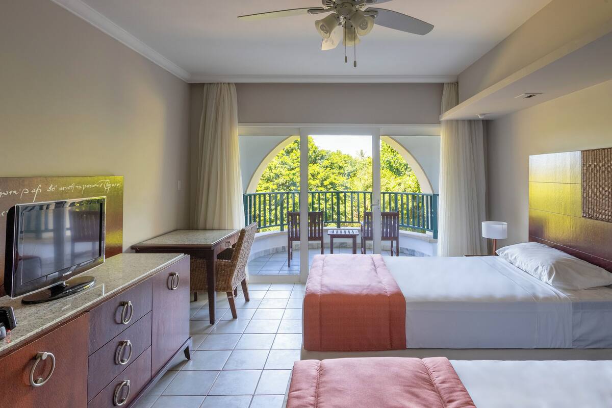 A hotel room with a bed, a desk with a TV, a balcony with a view of trees, and a ceiling fan.