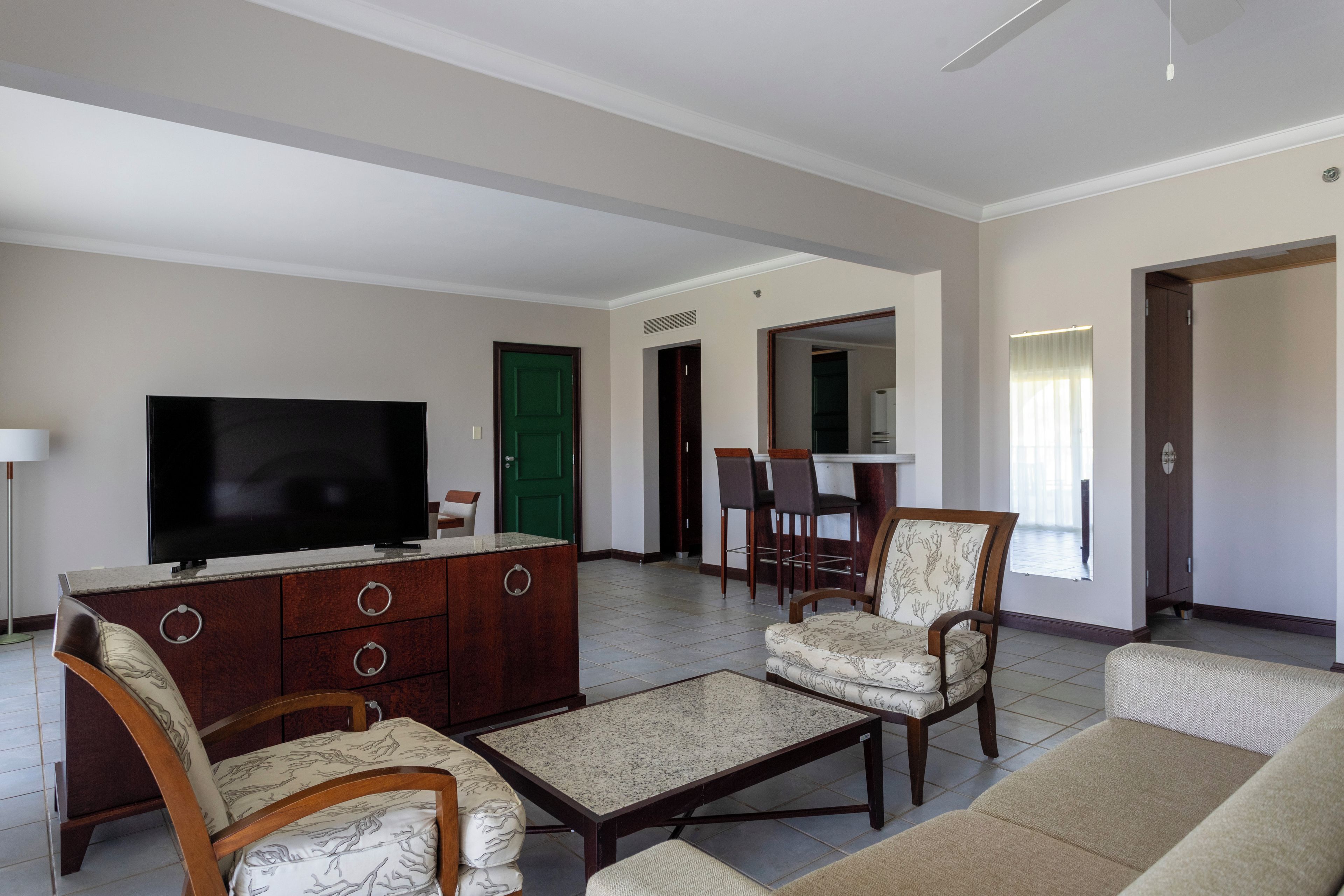 Uma sala de estar com televisão, uma área de jantar com cadeiras e uma cozinha com geladeira.
