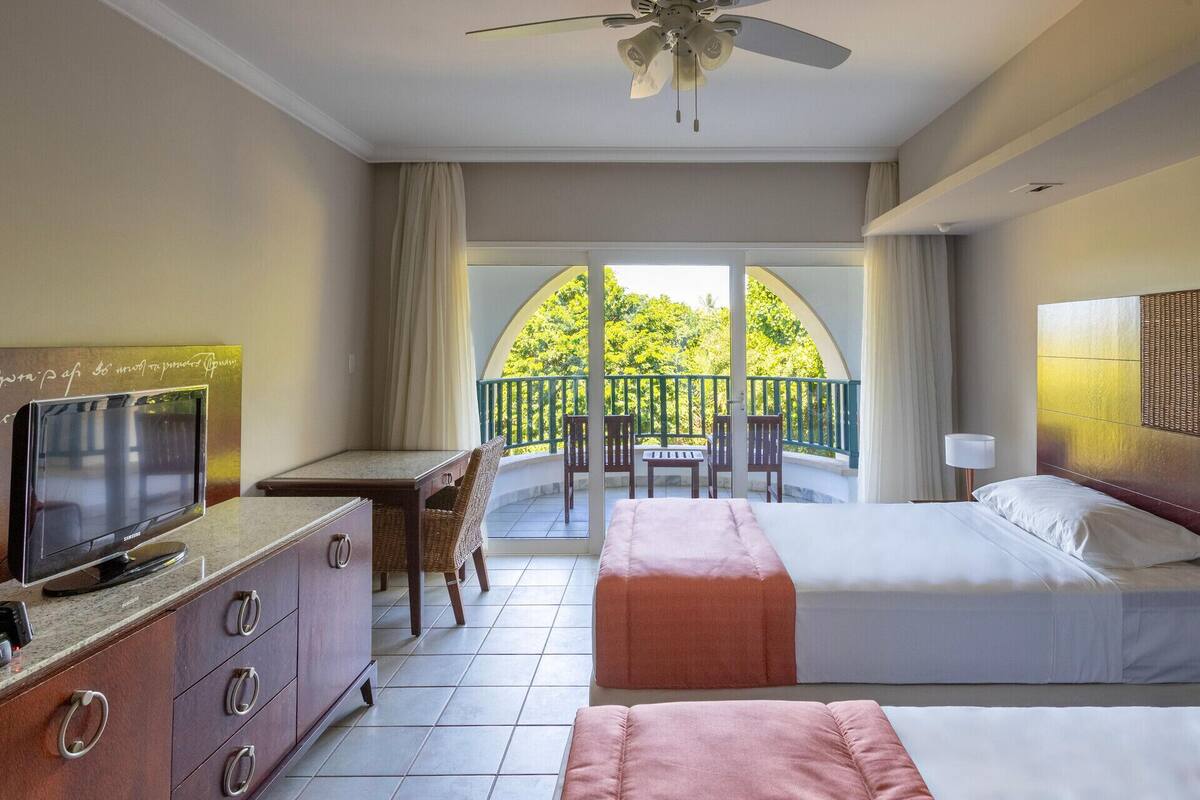 A hotel room with a bed, a desk with a TV, a balcony with chairs, and a ceiling fan.