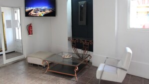 Un salon moderne avec une table basse en verre, un canapé et un fauteuil blanc. Un téléviseur à écran plat est fixé au mur, et on peut voir un extincteur.