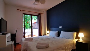 Une chambre d’hôtel avec un lit, deux lampes de chevet, un ventilateur de plafond, une télévision et une vue sur un balcon agrémenté d’une plante.