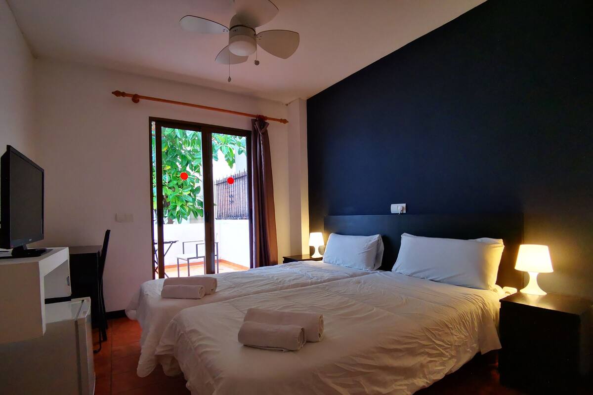 Ein Hotelzimmer mit Bett, zwei Nachttischlampen, Deckenventilator, Fernseher und Blick auf einen Balkon mit Pflanze.