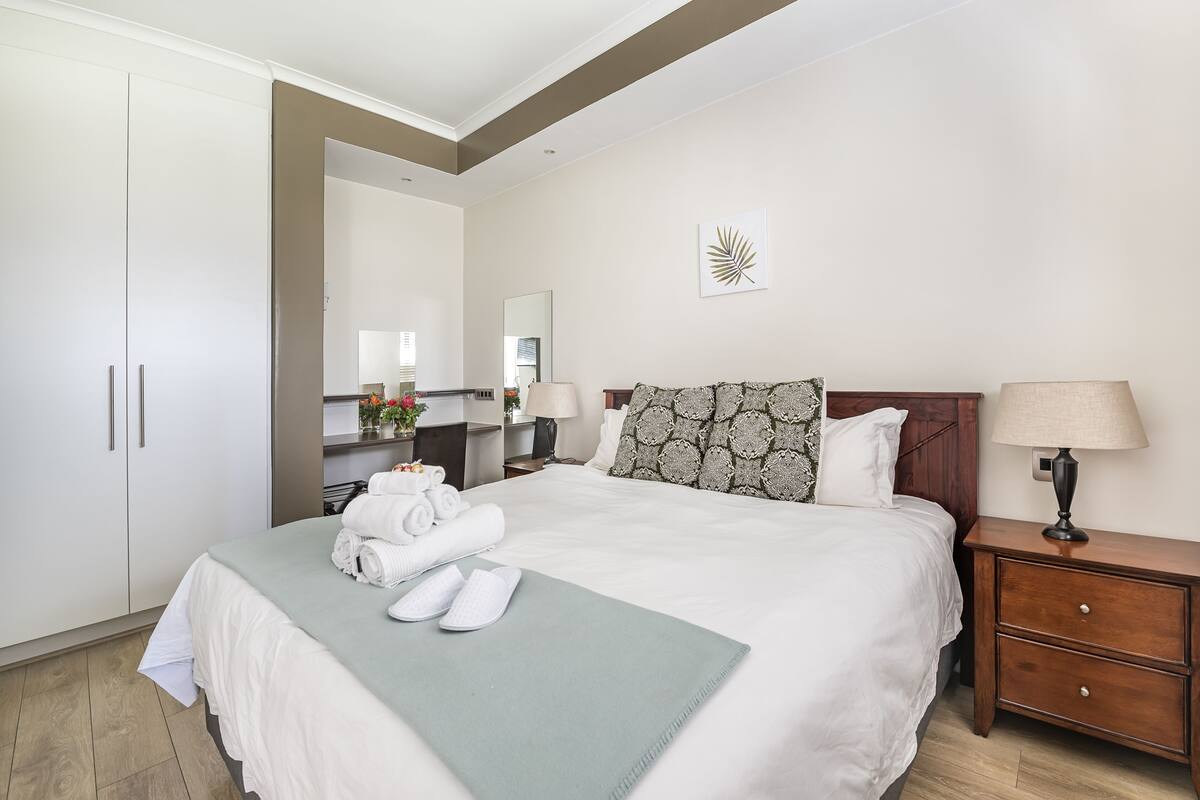 Una cama bien hecha con sábanas blancas, un plaid verde y toallas enrolladas. Hay una mesita de noche de madera con una lámpara y un espejo encima.