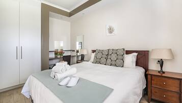 A neatly made bed with white linens, a green throw, and rolled towels. There is a wooden nightstand with a lamp and a mirror above it.