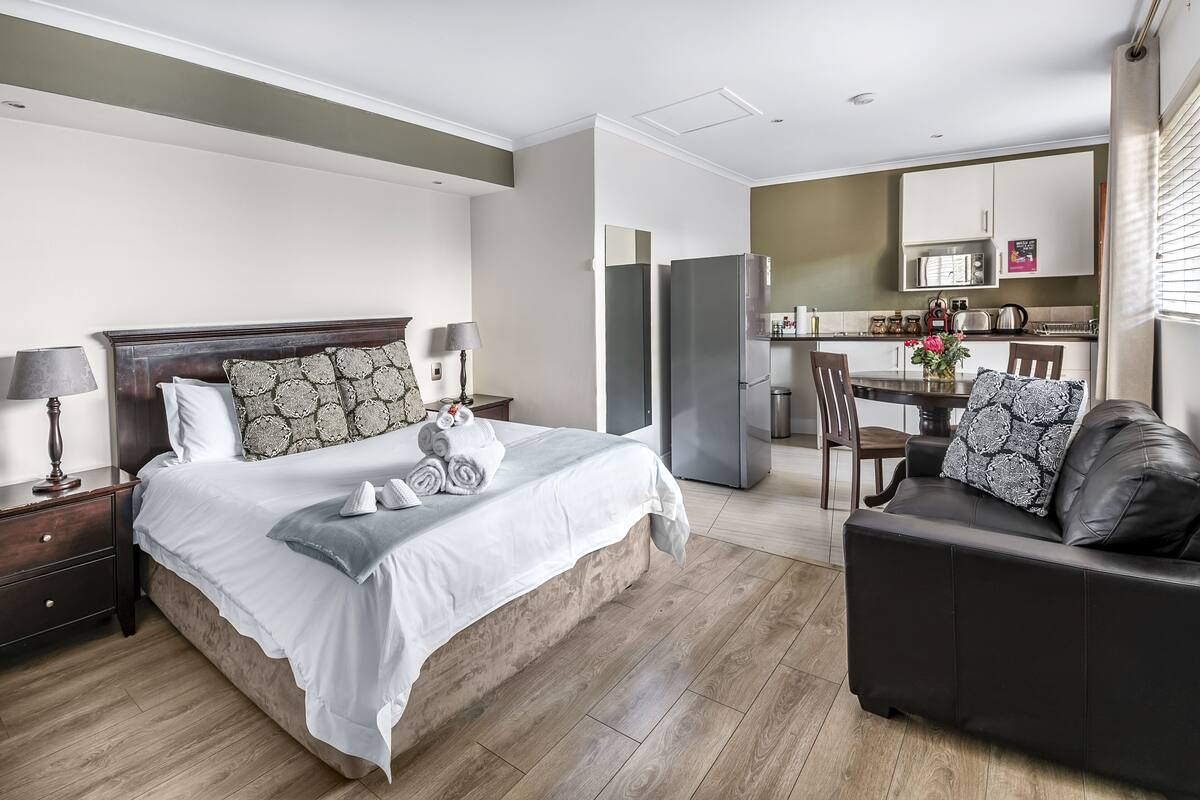 Una habitación de hotel moderna con una cama grande, un sofá de cuero oscuro, una mesa de comedor y una zona de cocina.