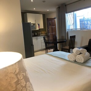 A hotel room with a bed, a table with towels, a dining area with chairs, a kitchen area, and a window with a view of buildings. - The Haystack on Homestead (Sandton)