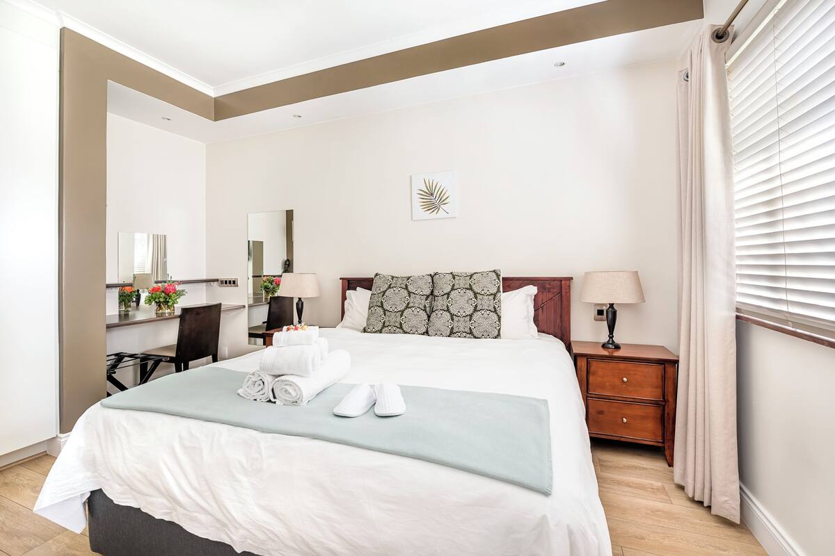 Una cama bien tendida con sábanas blancas y un plaid verde, una mesita de noche de madera con una lámpara, un espejo y una obra de arte colgada en la pared.