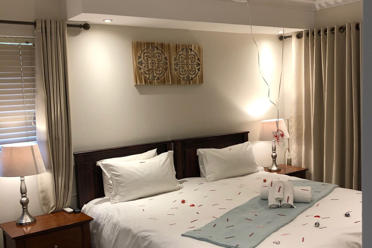 A neatly made bed with white linens, a bedside table with a lamp, and a wall-mounted artwork.