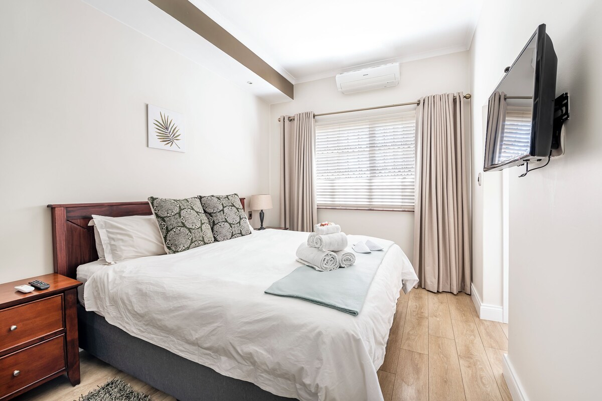 A neatly made bed with white linens, a bedside table with a lamp, and a wall-mounted TV.