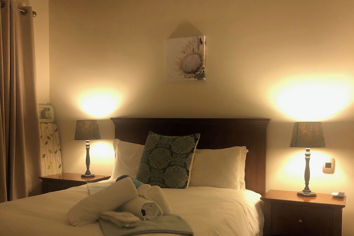 A hotel room with a bed, two bedside tables with lamps, a nightstand, a wall clock, and a decorative item above the bed.