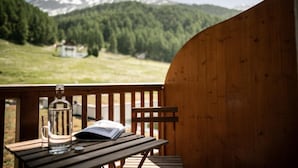 Una terraza de madera con una mesa, una botella y un vaso. Se divisa un paisaje montañoso al fondo.