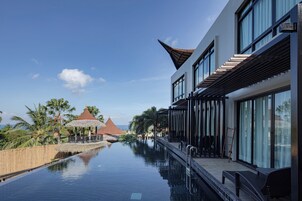 Ein modernes Hotel mit einem Infinity-Pool, einer überdachten Außenbereich und Blick auf Palmen sowie ein traditionelles Gebäude im Hintergrund.