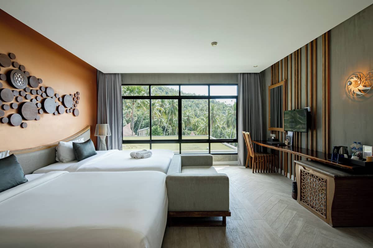 A modern hotel room with two beds, a large window with a view of greenery, a wooden desk, and a wall-mounted TV.
