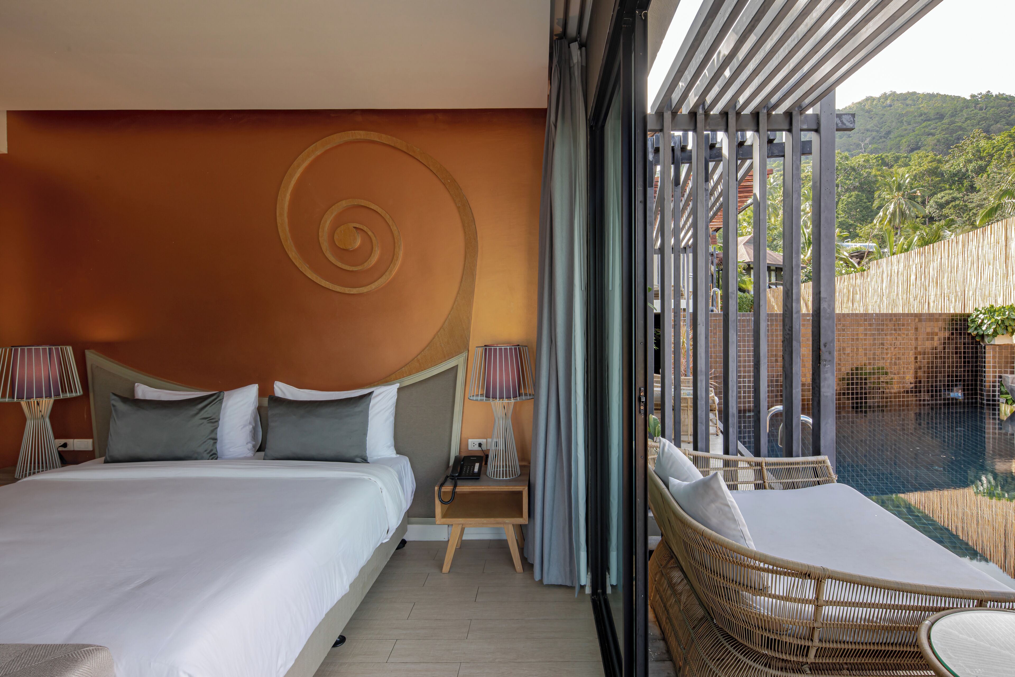 A hotel room with a large bed, a bedside table with a lamp, a small table with a chair, and a view of a pool and greenery.