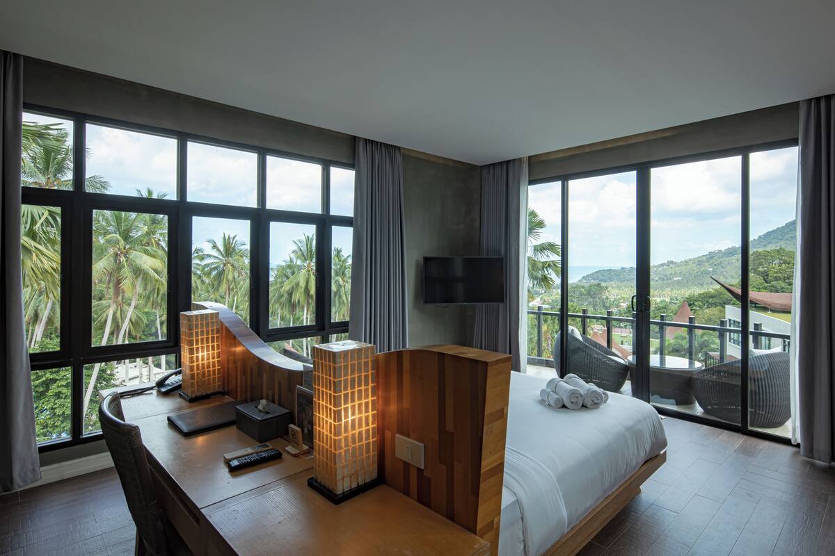 A modern hotel room with a large bed, a desk with a lamp, a TV, and a balcony with a view of palm trees and hills.