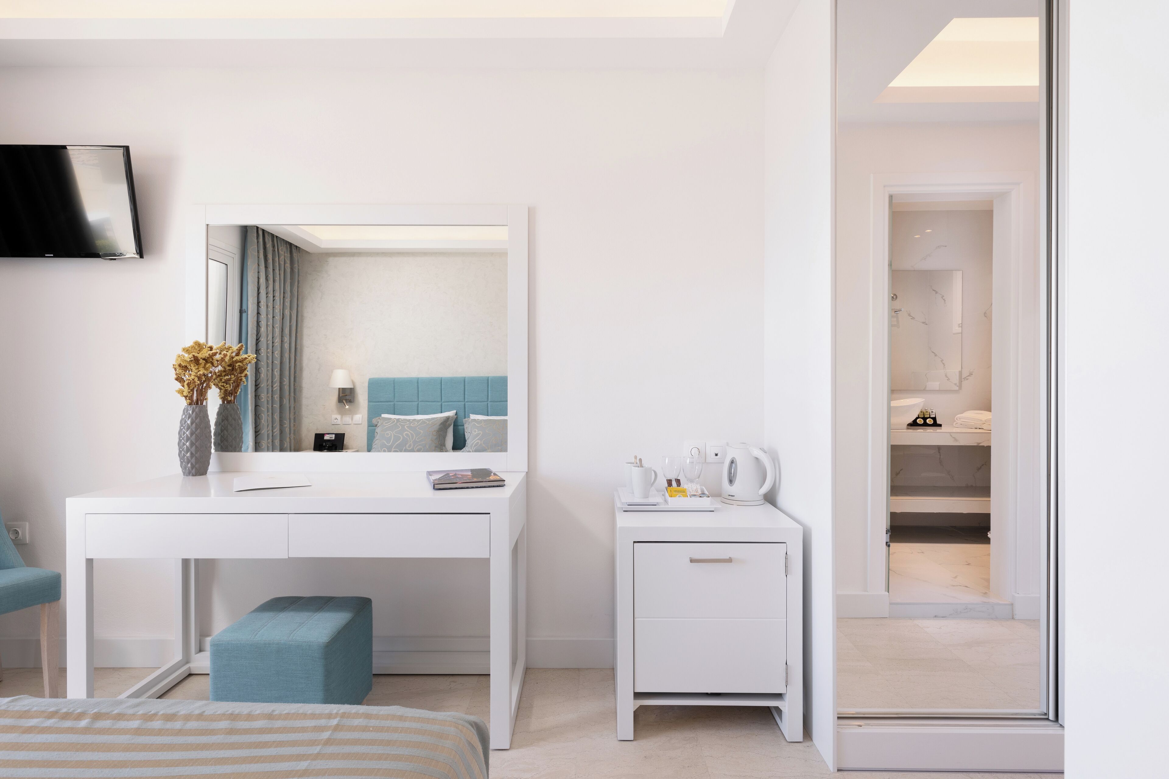 A modern hotel room with a white desk, a small bedside table, a blue chair, and a large mirror.