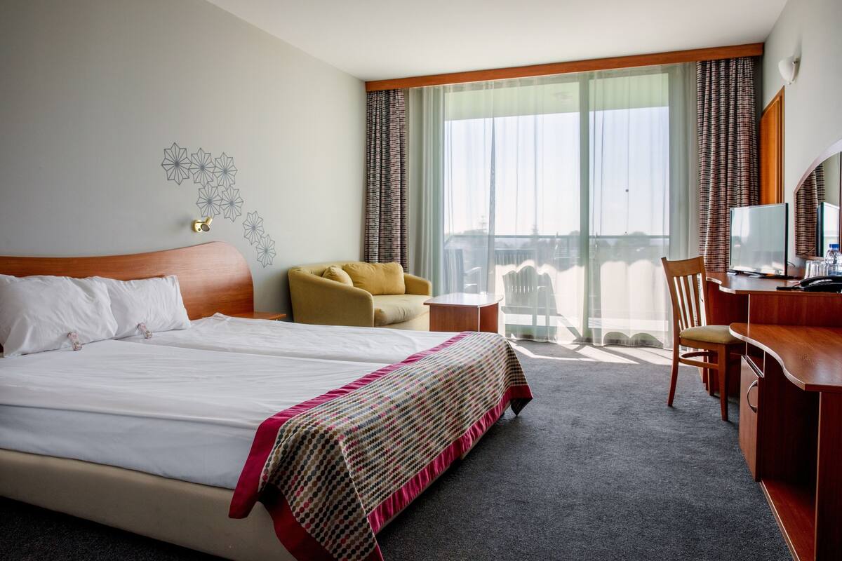 A hotel room with a large bed, a desk, a chair, and a view of the city through large windows.