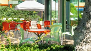 An outdoor seating area with tables and chairs, a white umbrella, and a view of a modern building with large windows. - MPM Kalina Garden Hotel (Sunny Beach)