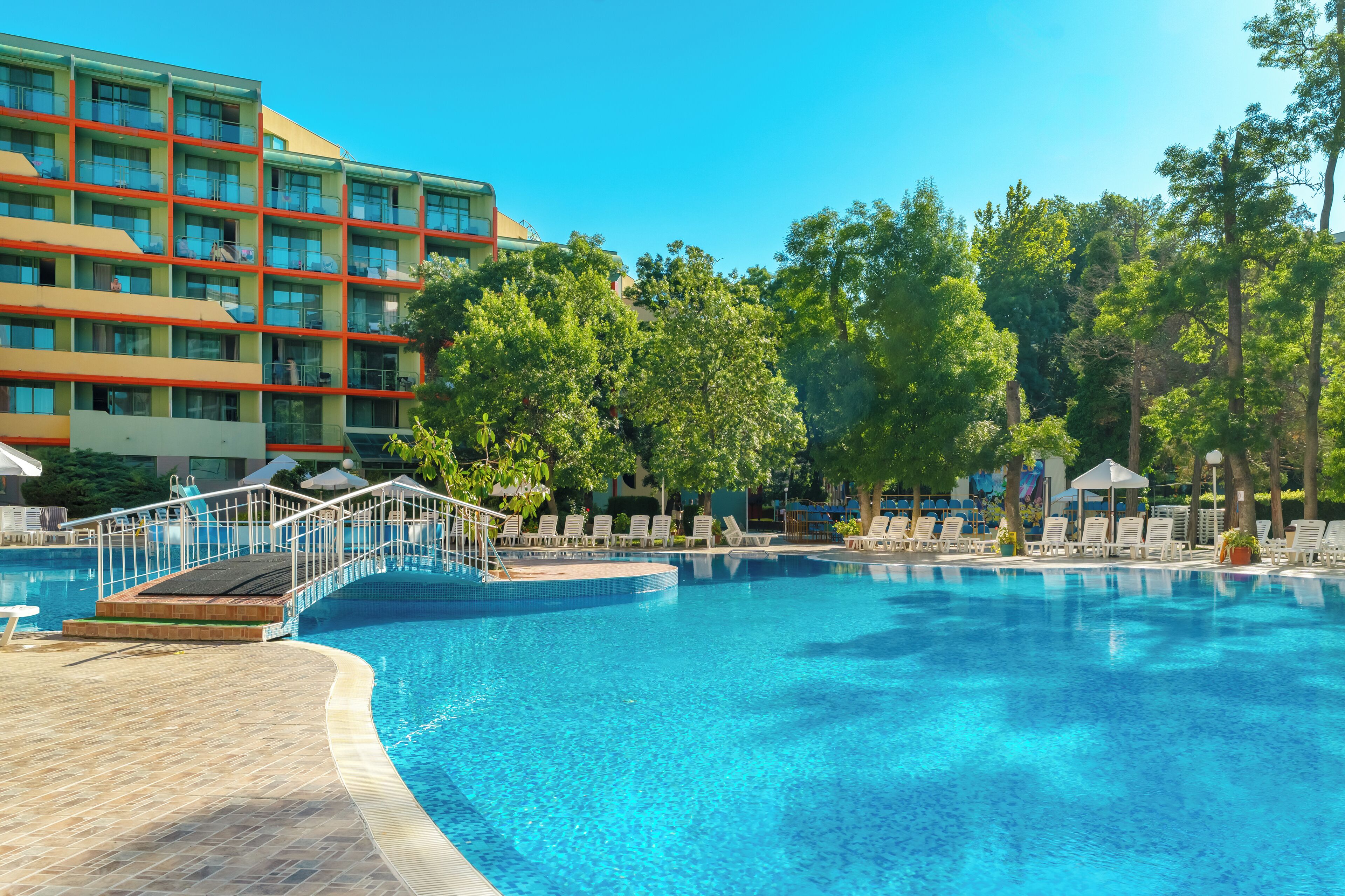 A swimming pool with lounge chairs and a bridge, surrounded by trees and a hotel building.