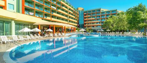Un hotel con una piscina grande rodeada de tumbonas y sombrillas, varios pisos con balcones y un cielo azul despejado.