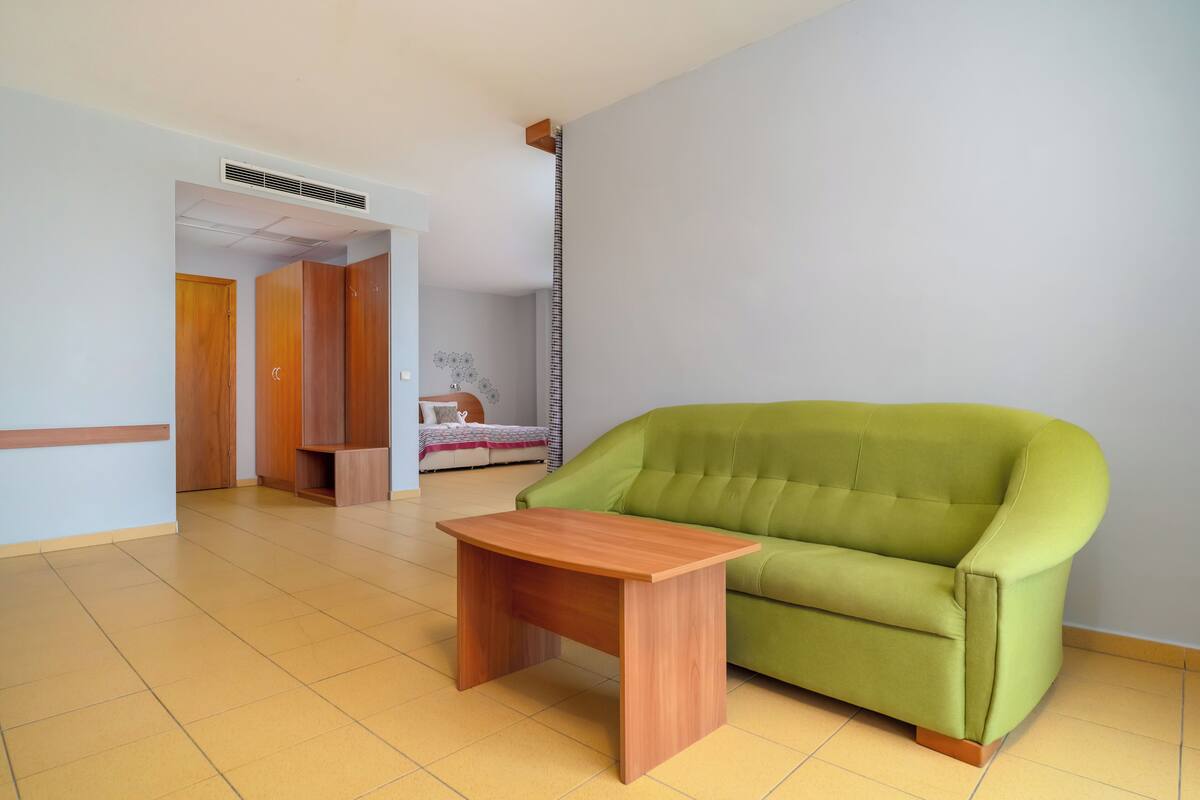 A room with a green sofa, a wooden table, and a bed with a white headboard.