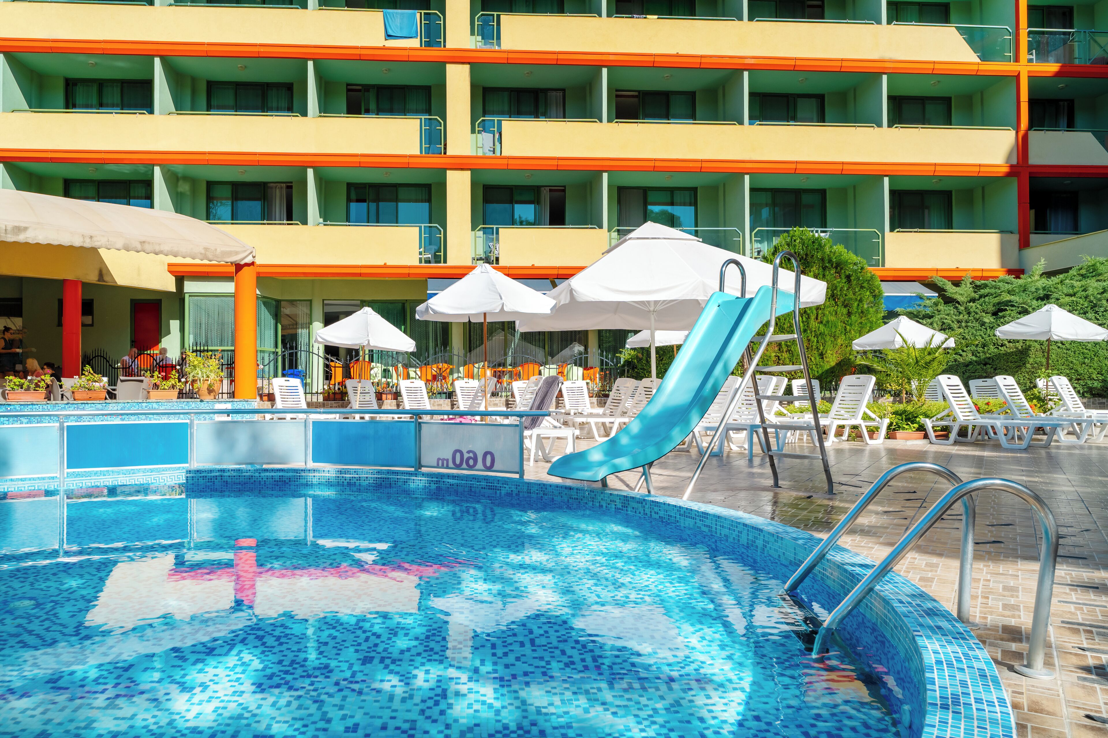 A swimming pool with a slide, lounge chairs, umbrellas, and a multi-story building with balconies.