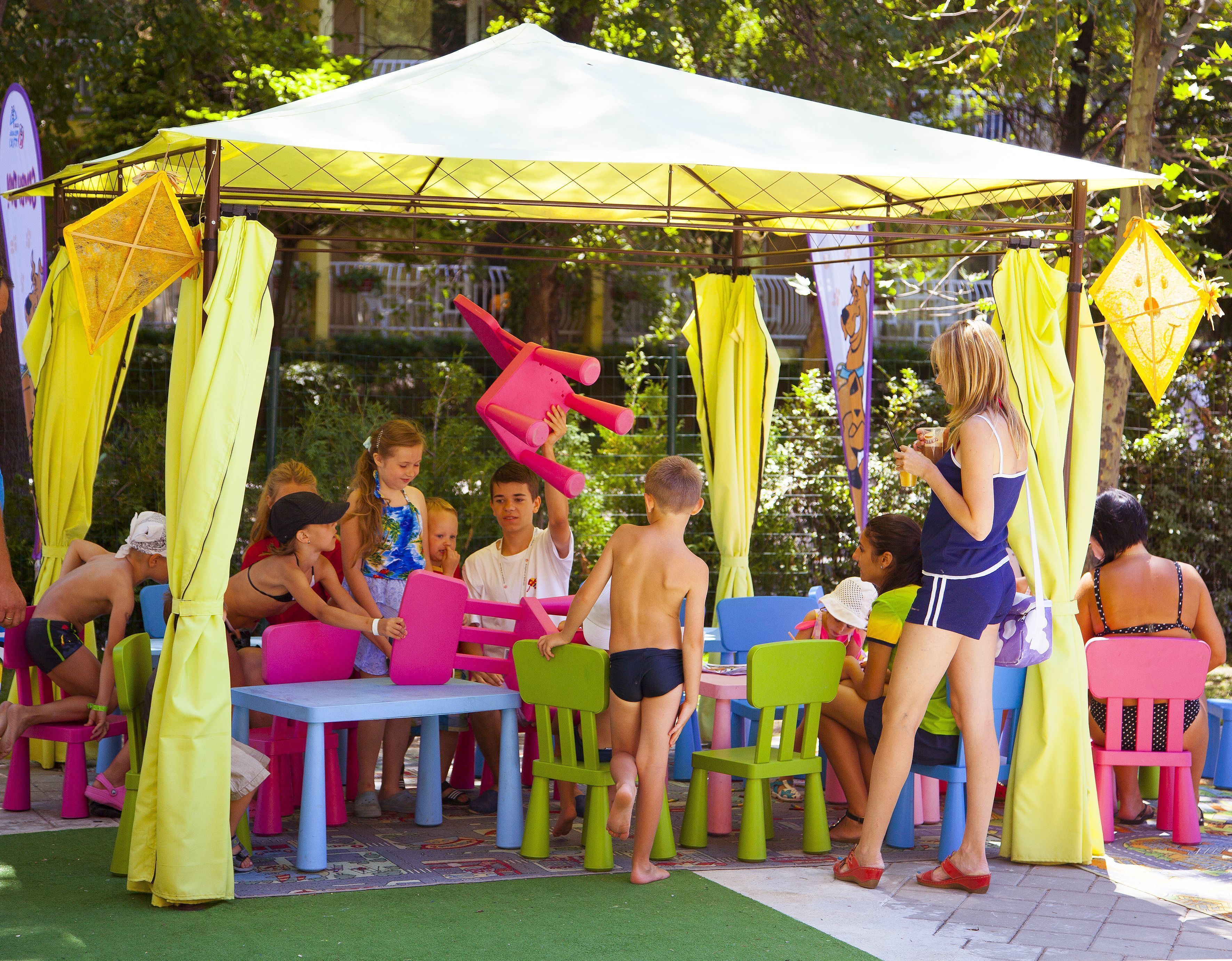 A covered outdoor seating area with colorful plastic chairs and tables, surrounded by greenery.