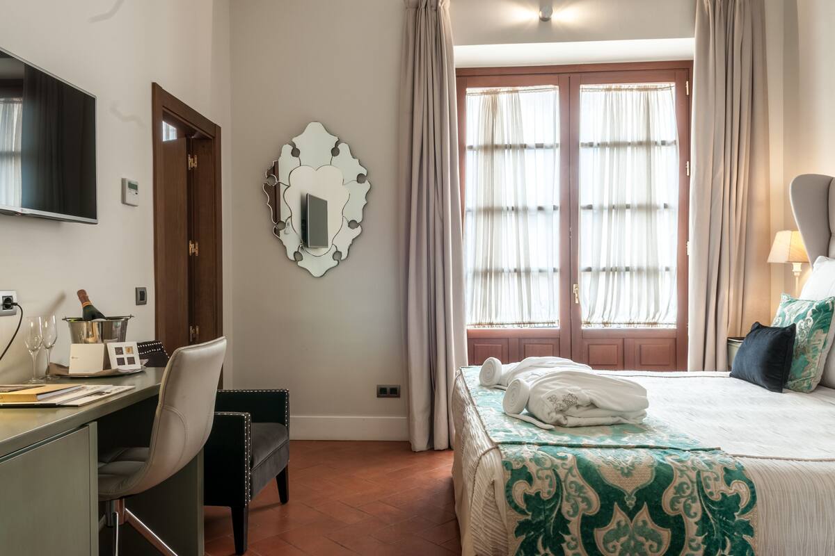 Ein Hotelzimmer mit einem Bett, einem Schreibtisch mit Computer, einem Fernseher und einem Fenster mit Vorhängen.
