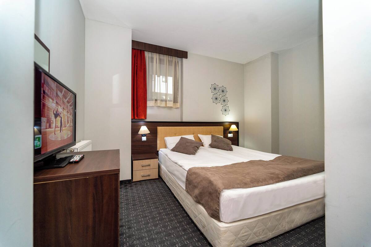 A hotel room with a bed, bedside tables, a TV, and a window with curtains.